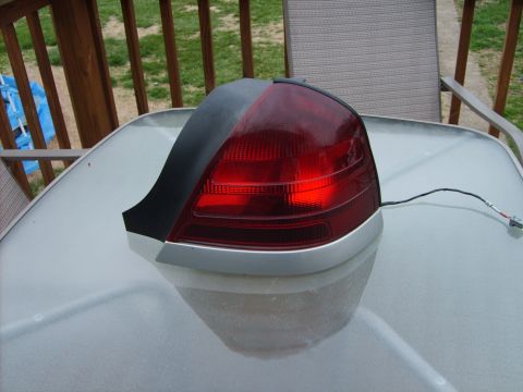 1993 Ford Crown Victoria - A removed rear taillight assembly with a red lens, black upper trim section, silver lower molding and a bare bulb-socket pigtail, set on a…