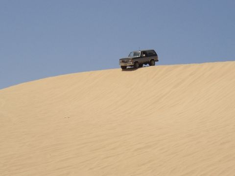 1989 Ford Bronco - A dark SUV navigates a sandy incline on a bright, clear day.