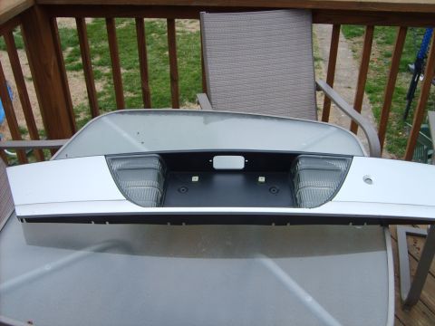 1993 Ford Crown Victoria - A white rear deck lid / trunk trim panel with an integrated black license plate recess and clear backup-light lenses, propped upright on a…