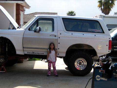 1996 Ford Bronco - Side view of a white Ford Bronco desert-race build in a driveway, hood up and front axle on jack stands with the wheels off, riveted fiberg…