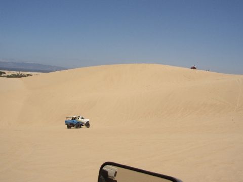 1989 Ford Bronco - A two-tone pickup truck navigates sandy terrain under a clear blue sky, with a watermark indicating SUPERMOTORS.