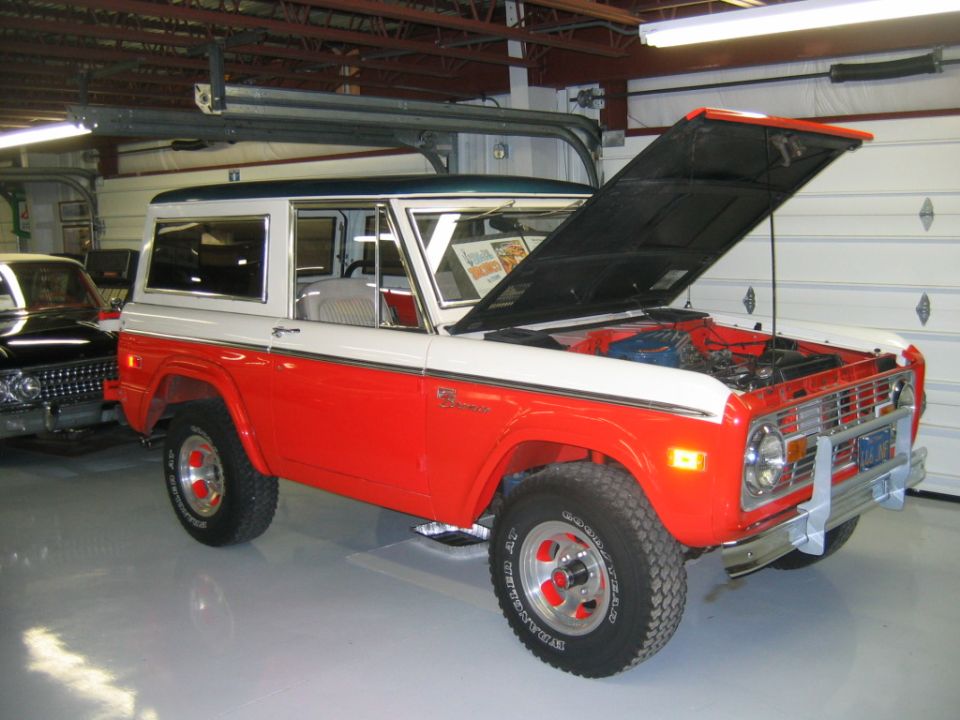 Red and white Ford Bronco with hood open, parked indoors next to a black car
