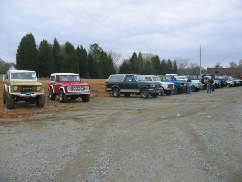 1992 Ford Mustang - A collection of Ford Broncos and other SUVs are parked in a gravel lot with trees in the background.