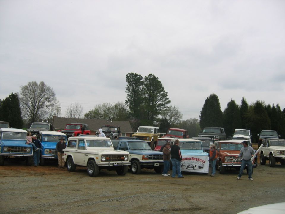 Group of Ford Broncos and trucks parked outside, some people milling around