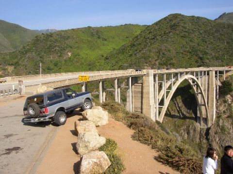 1989 Ford Bronco - A two-tone blue and gray Ford Bronco is parked on the side of the road overlooking a large bridge.