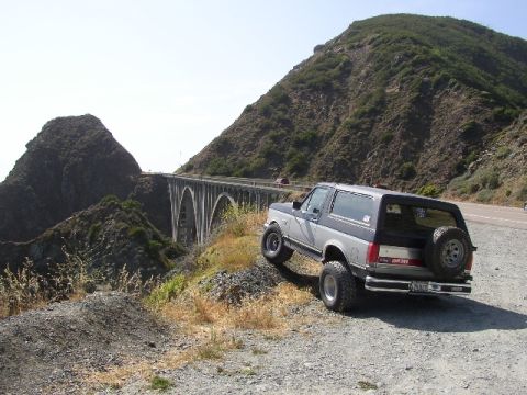 1989 Ford Bronco - A 1989 Ford Bronco is parked on a dirt embankment overlooking a scenic coastal road and bridge.