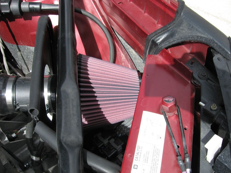 Red engine bay with a pink K&N air filter and black hoses. A white sticker with text is visible