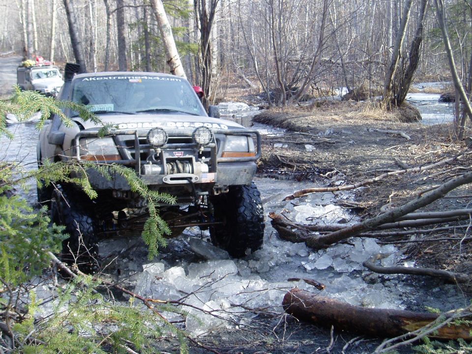 Off-road truck with winch and lights driving through icy stream. Another vehicle is visible behind it