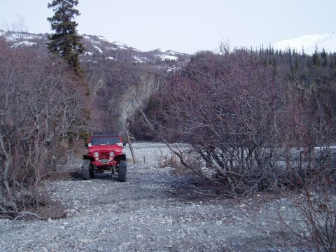 2000 Ford E350 - Red Jeep CJ-5 parked on a gravelly riverbed with bare trees and snow-capped mountains in the background.