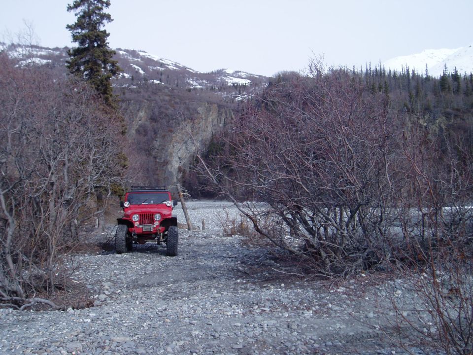 Red Jeep CJ-5 parked on a gravelly riverbed with bare trees and snow-capped mountains in the background