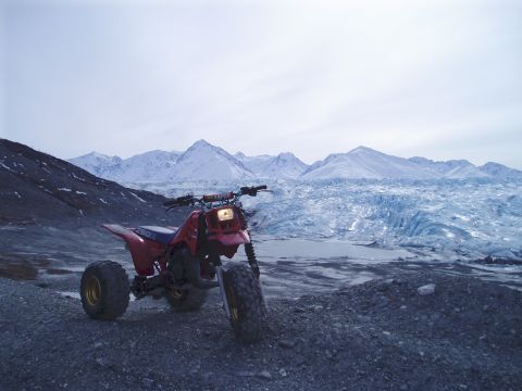 2000 Ford E350 - A red three-wheeled ATV sits in a desolate landscape, with a vast glacier and imposing mountains dominating the horizon.