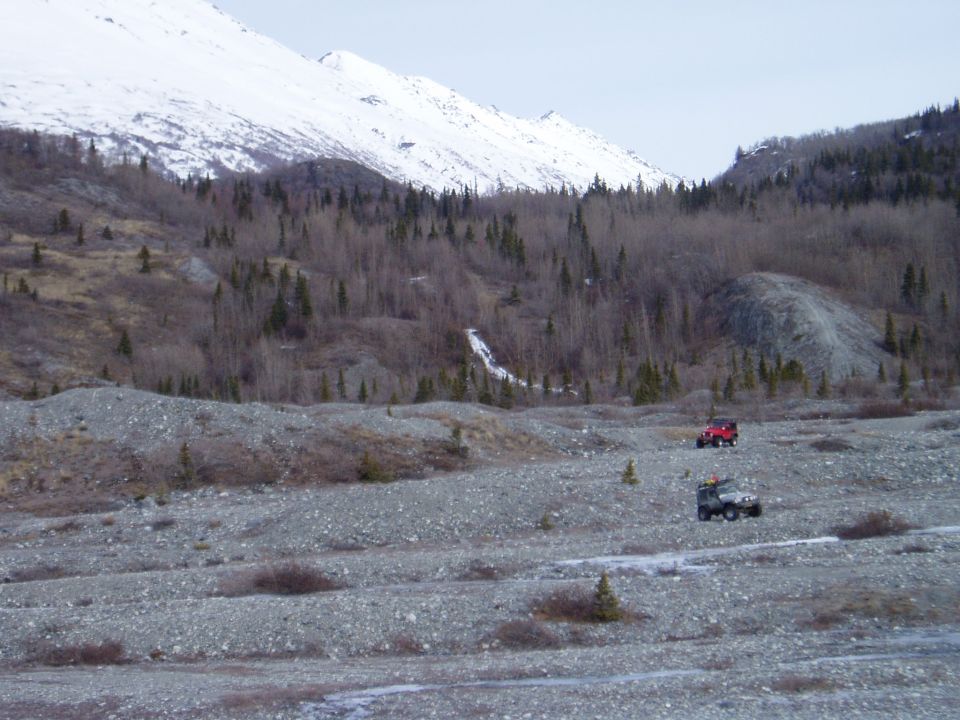 Two off-road vehicles drive through a rocky, mountainous terrain with snow-capped peaks in the background