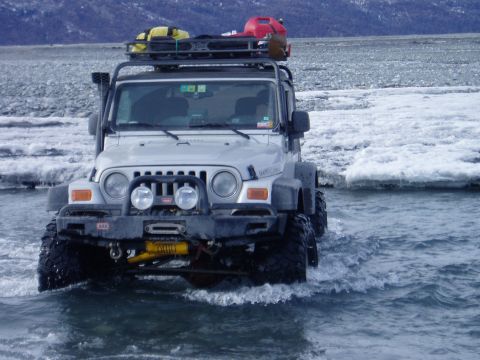 2000 Ford E350 - A silver Jeep Wrangler navigates through a shallow, icy river, equipped with a roof rack carrying supplies for an expedition.