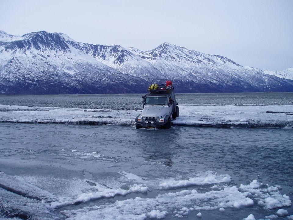 Silver Jeep Wrangler with roof rack crossing shallow icy water, snow-covered mountains in background