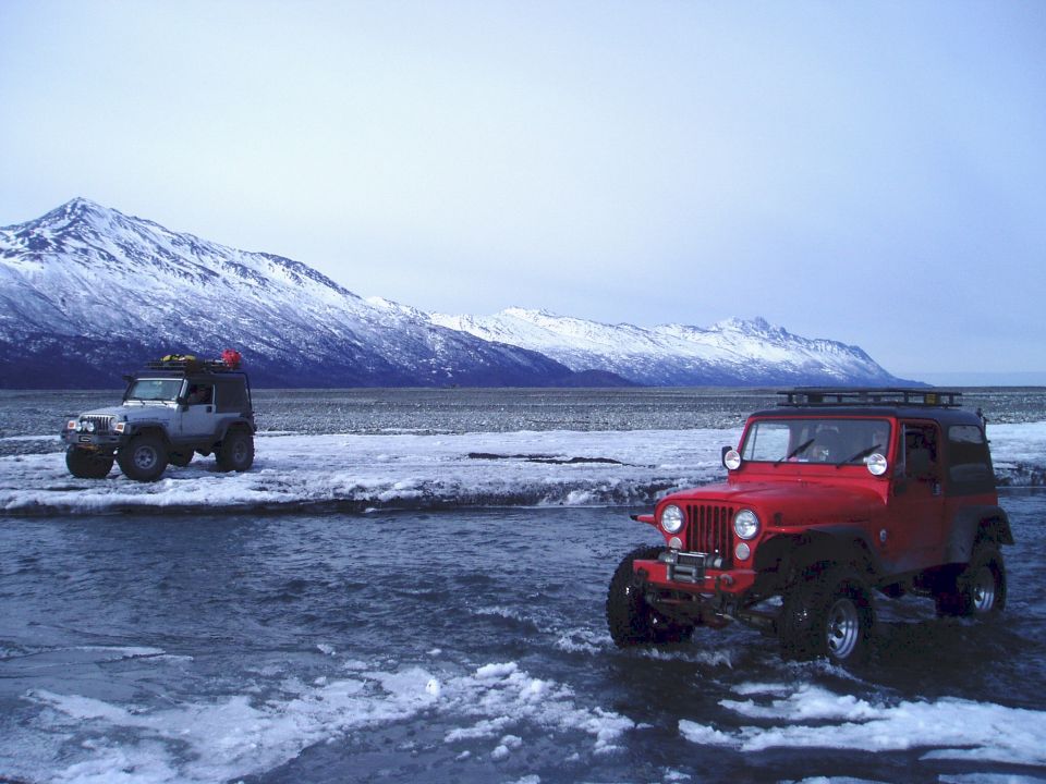 Red Jeep Wrangler and a silver Jeep Cherokee drive through icy water with snowy mountains behind them