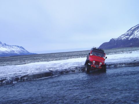 2000 Ford E350 - A red jeep navigates a frozen riverbed under a cloudy sky with snow-capped mountains in the distance.