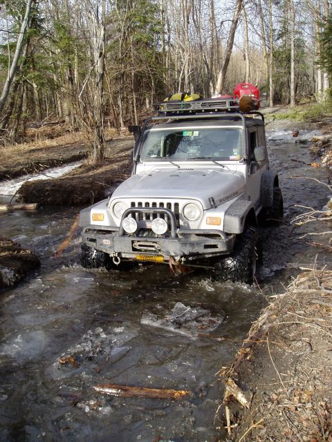 2000 Ford E350 - A Jeep Wrangler navigates a partially frozen stream surrounded by trees, with a roof rack loaded with equipment.