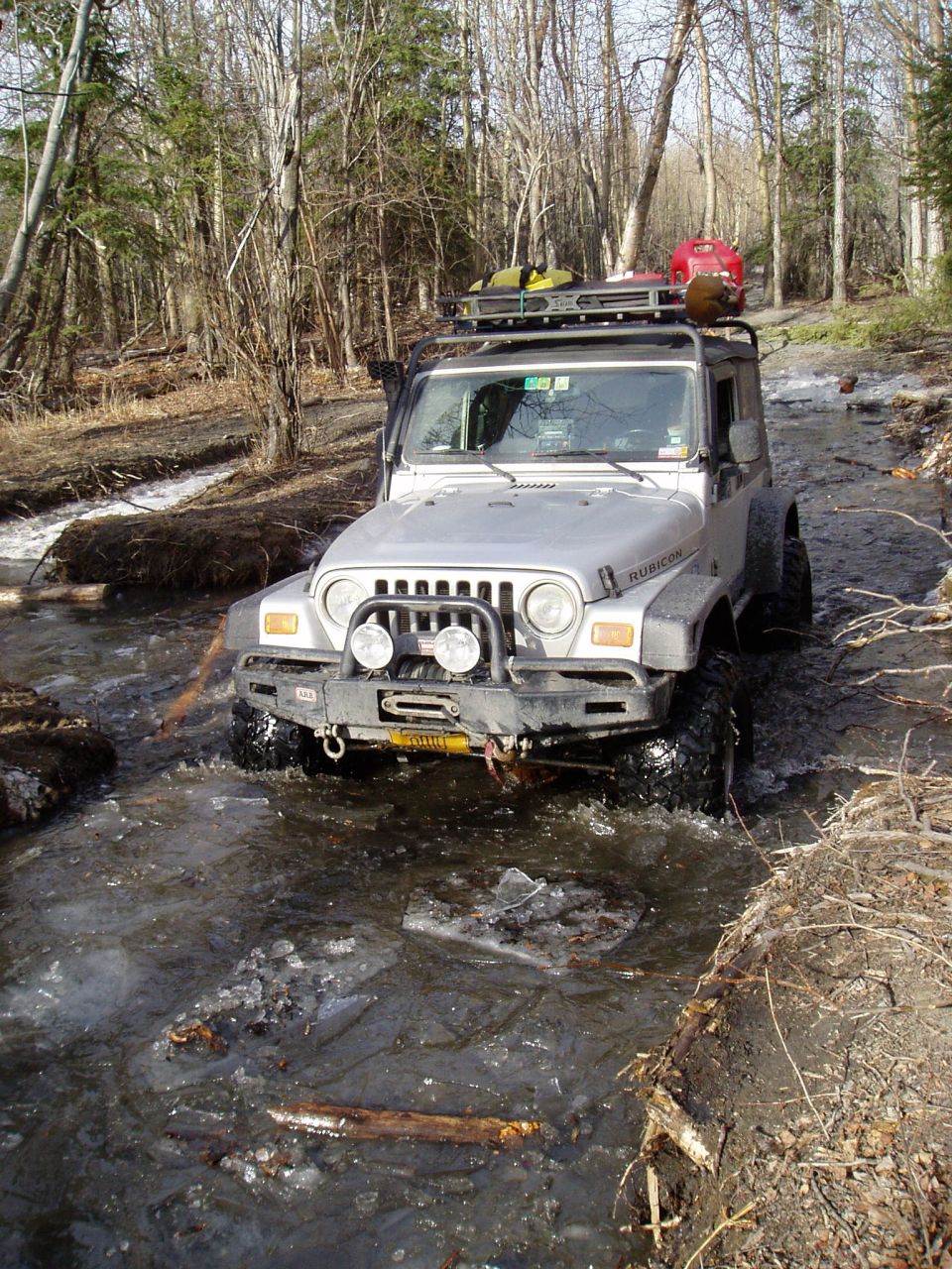 Silver Jeep Wrangler driving through a shallow, icy stream in a wooded area. It has a roof rack with gear