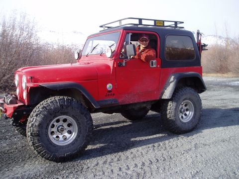 2000 Ford E350 - A red Jeep Wrangler with a black hardtop and roof rack is parked on a gravel surface with a driver inside.
