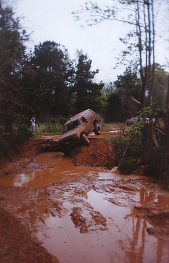 1984 Ford Bronco - Off-road vehicle struggling to navigate a treacherous mud pit on a wooded trail.