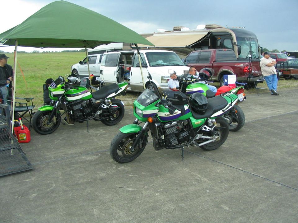 Two green Kawasaki motorcycles parked under a canopy, with vans and an RV in the background