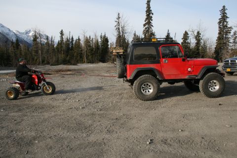 2003 Jeep TJ - A red Jeep Wrangler is being towed by a red ATV in a gravelly area with trees and mountains in the distance.