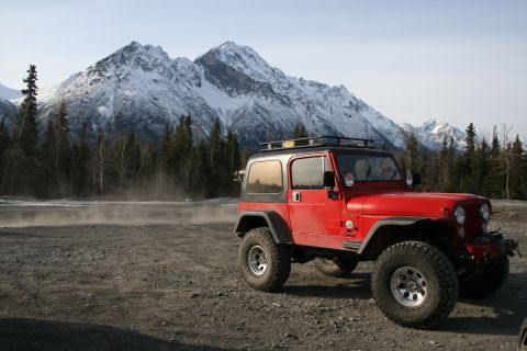 2003 Jeep TJ - A red Jeep Wrangler sits parked on a gravel lot with snow-covered mountains and pine trees in the background.