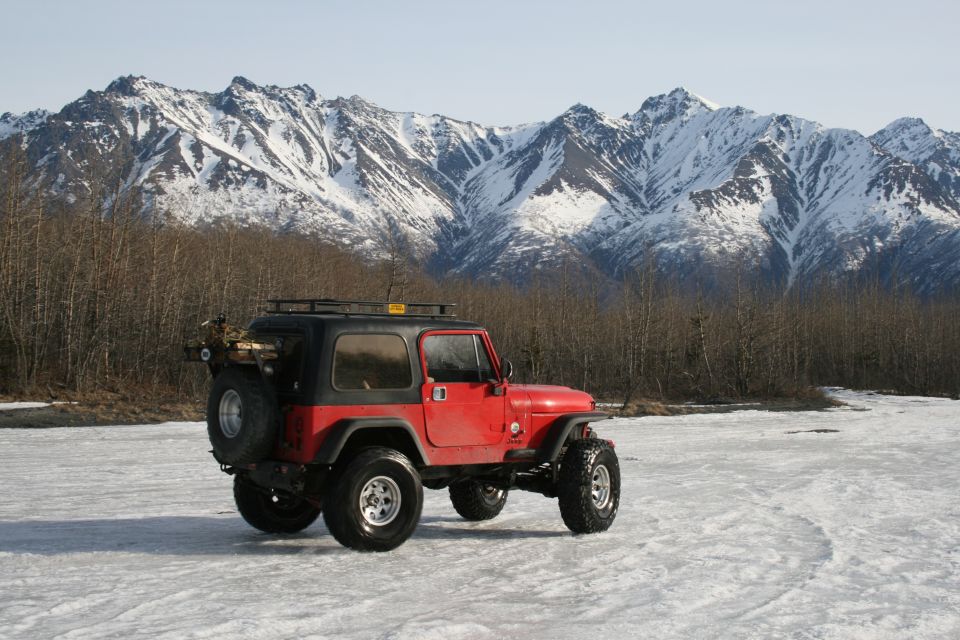 Red Jeep TJ with a black hardtop and roof rack parked on ice, with snow-covered mountains in the background