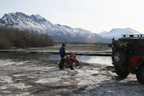 2003 Jeep TJ - A person stands by a red ATV next to a Jeep with a spare tire on the back, with snowy mountains visible.