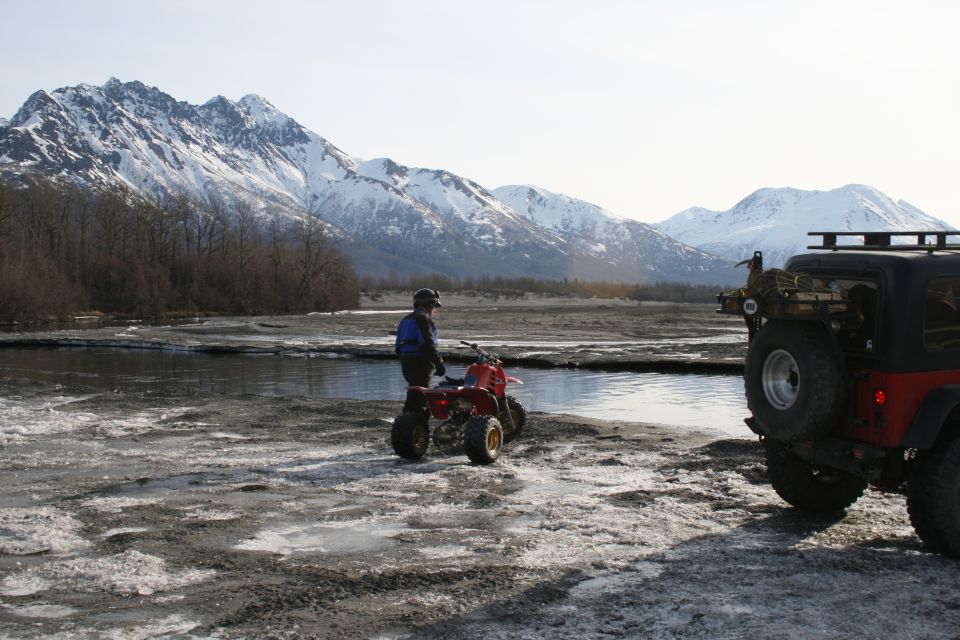 Red ATV and black Jeep parked on icy ground near a river with snow-capped mountains in the background