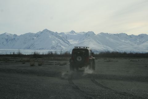 2003 Jeep TJ - A red Jeep Wrangler kicks up dust as it drives across a barren landscape towards a , snow-covered mountain range.