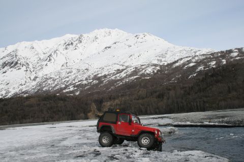 2003 Jeep TJ - A red 2003 Jeep TJ sits by a flowing river with a snow-capped mountain range behind it.