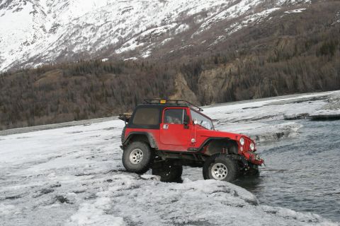 2003 Jeep TJ - A red 2003 Jeep TJ navigates icy terrain near a body of water, with a snow-covered mountain range in the background.
