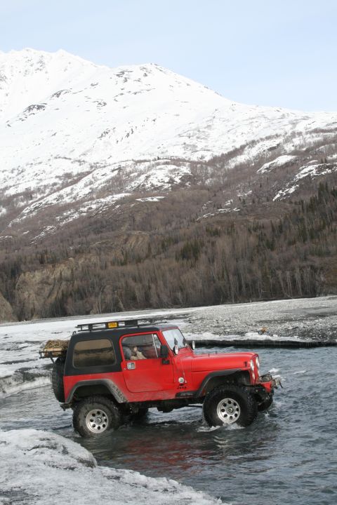 2003 Jeep TJ - A red Jeep Wrangler navigates a shallow riverbed with a snow-capped mountain range behind it.