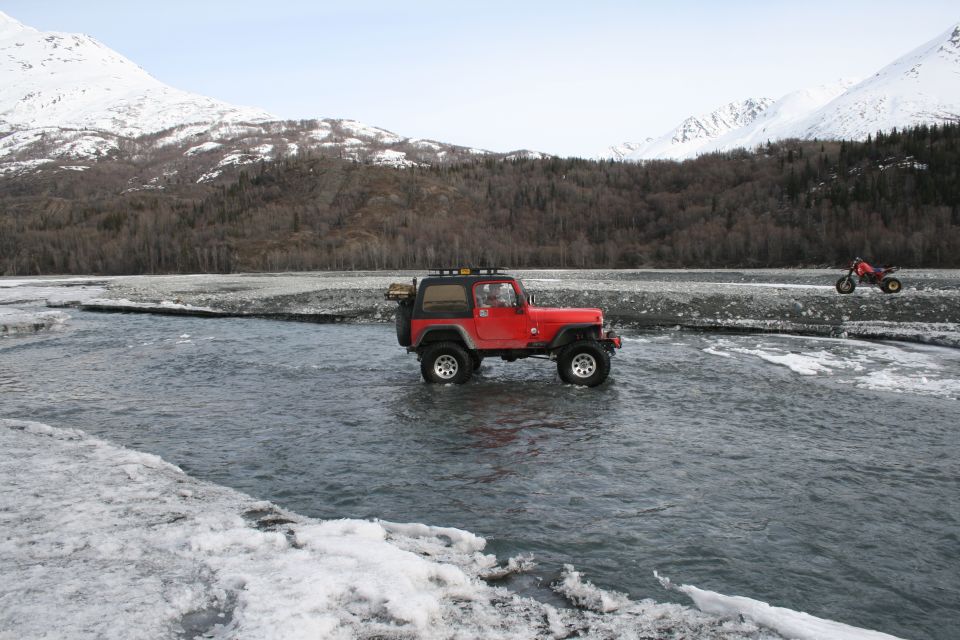 Red Jeep Wrangler driving through shallow water with snow-capped mountains in background