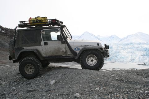 2003 Jeep TJ - A white 2003 Jeep Wrangler Rubicon, equipped for off-road adventure with a snorkel and roof rack, is parked on a gravelly terrain with a gl…