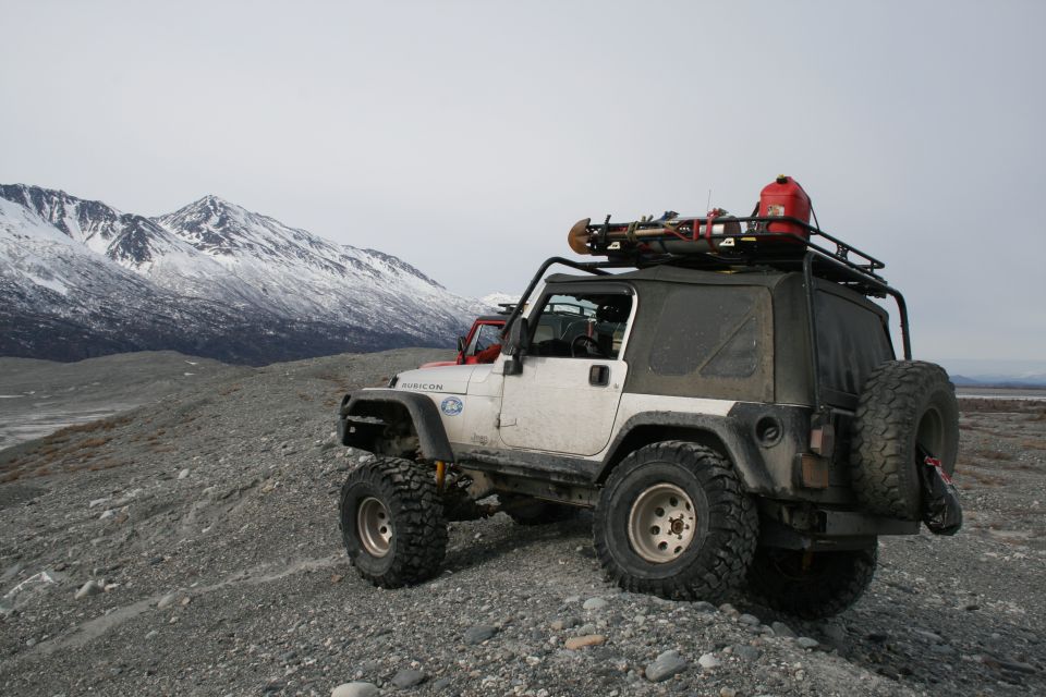 White Jeep Wrangler with a roof rack carrying a red gas can and gear, parked on a gravelly terrain with snow-capped mountains in the background