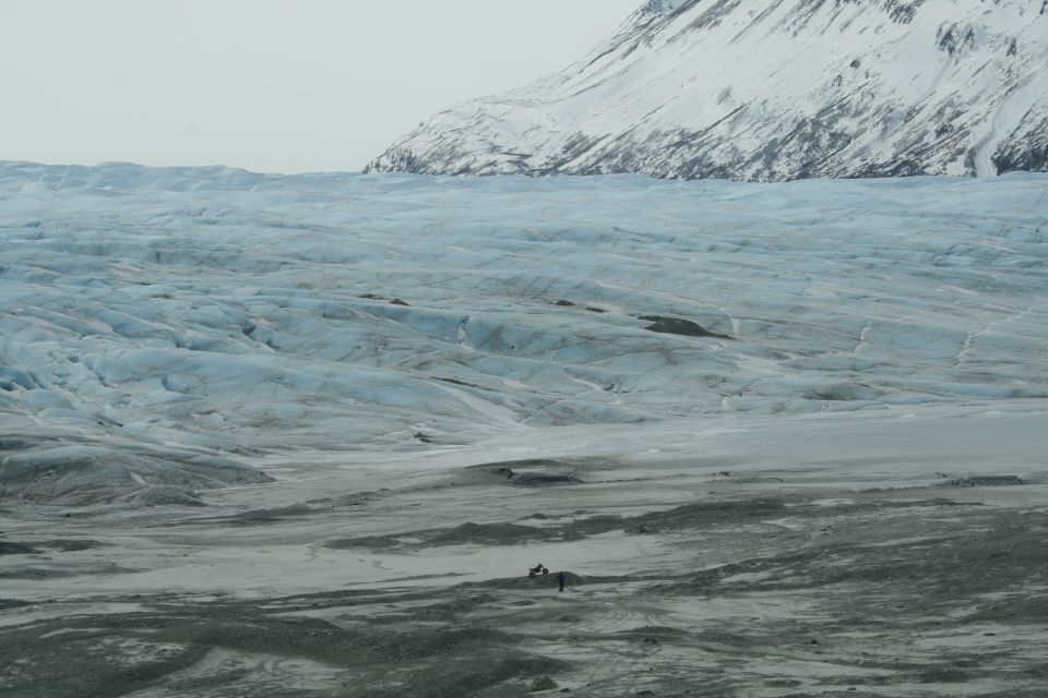 Expansive glacier with blue ice formations, leading to a barren, rocky foreground and a snow-covered mountain peak under a cloudy sky