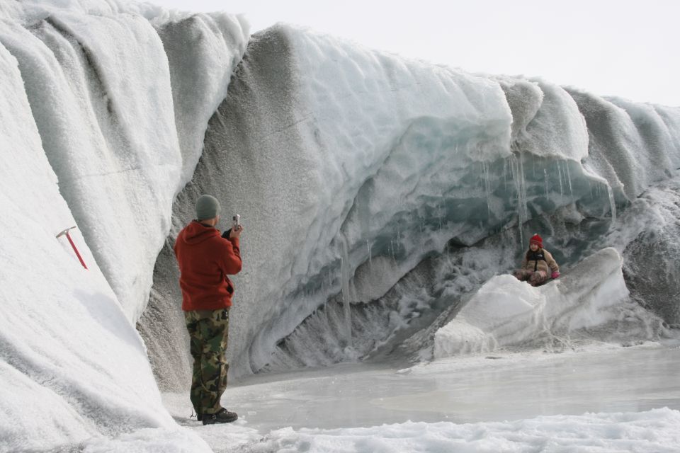 Two people stand on a glacier, one taking a photo, the other sitting on ice