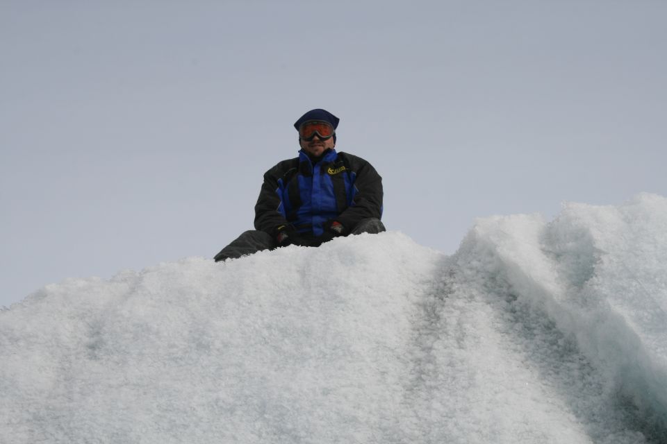 Person wearing winter clothing and goggles sits on a large ice formation under a clear sky