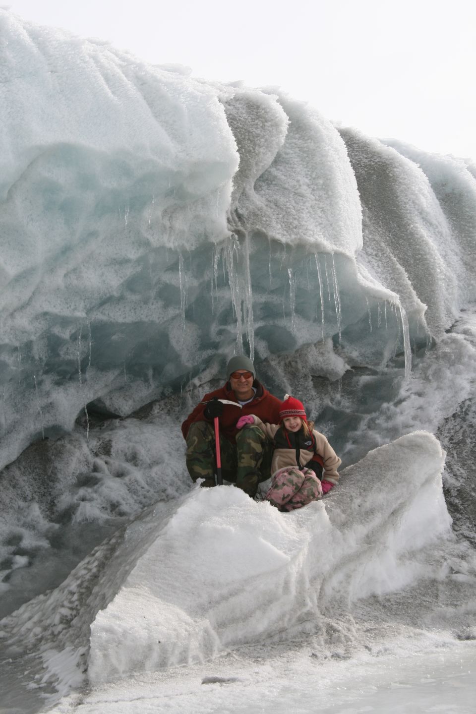 Adult and child sit on ice formations in front of a glacier