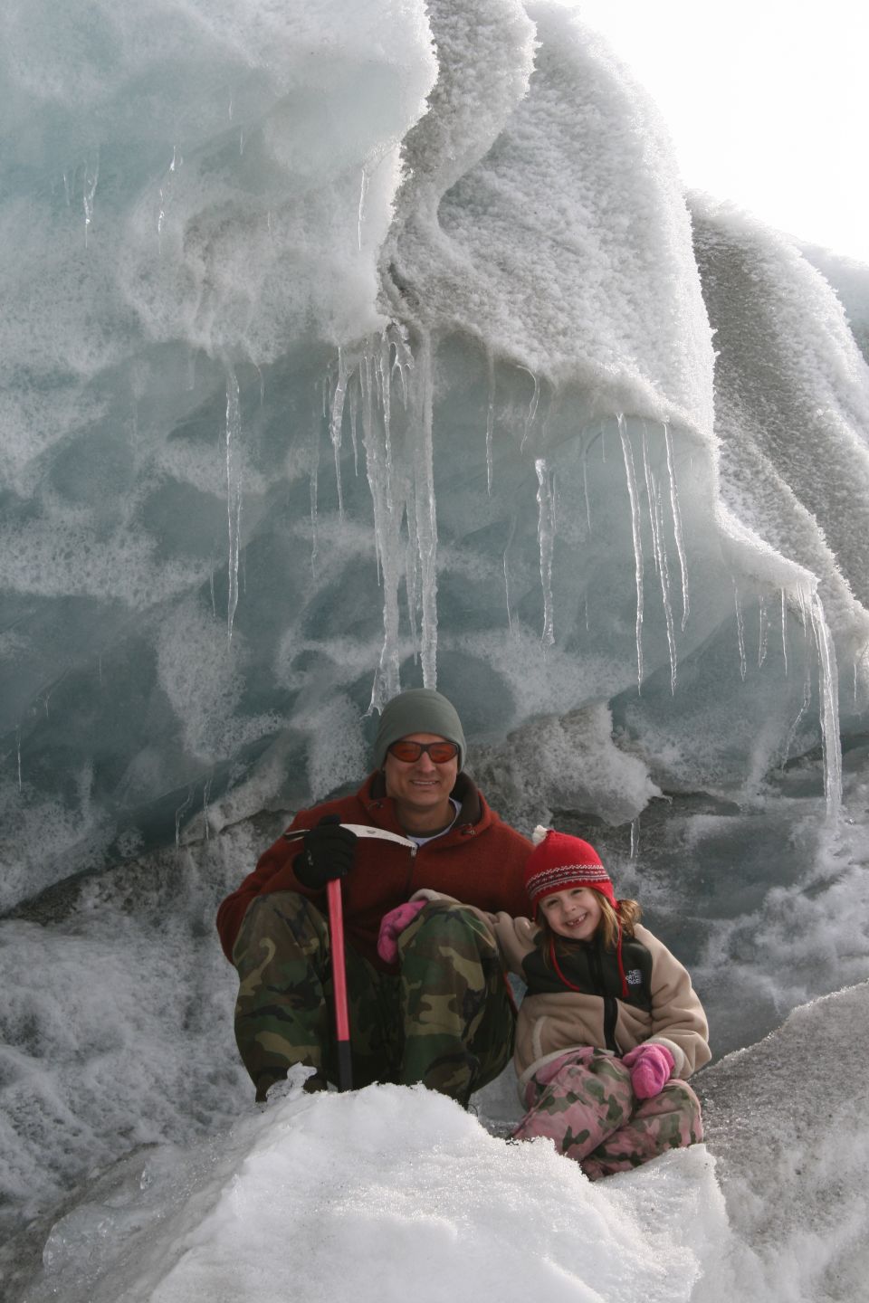 Man and child in winter clothing sit on snow in front of a glacial ice wall with icicles