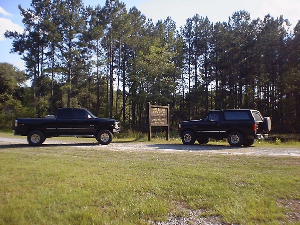 Black Ford pickup and black two-door SUV parked on gravel near pine trees and a wooden sign.