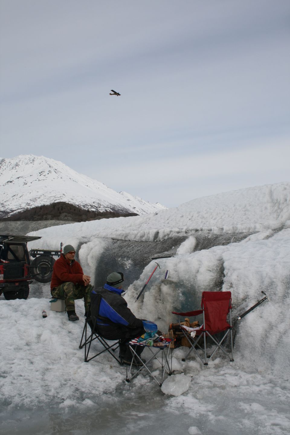 Two people sit in camping chairs near a glacier, with a small airplane flying in the distance
