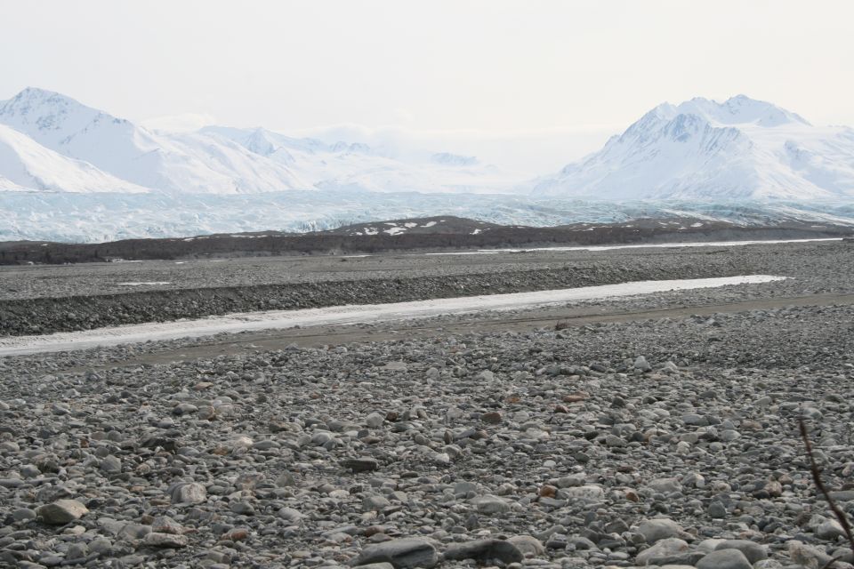 Snow-covered mountains and a vast glacier stretch across the horizon above a rocky plain with a stream