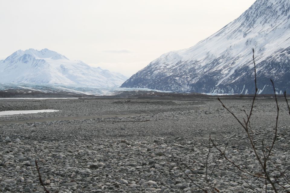 Rocky valley floor with a riverbed, leading to a vast glacier between snow-capped mountains under a hazy sky