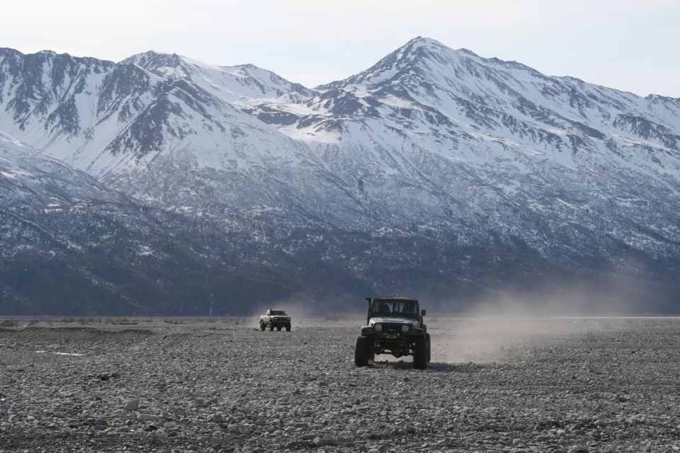 2003 Jeep TJ and another SUV driving on a rocky plain, kicking up dust, with snow-covered mountains behind them