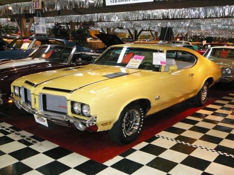 1997 Mercury Grand Marquis - A yellow classic car is displayed indoors with a checkered floor and other vehicles nearby.
