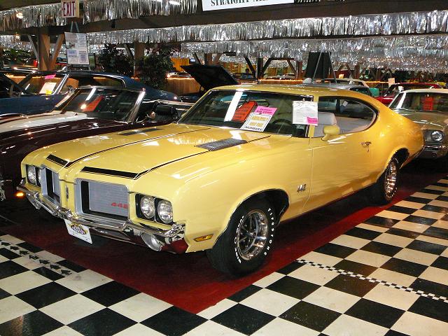 Yellow 1970s muscle car with black stripes on hood and grille. Other cars visible in background