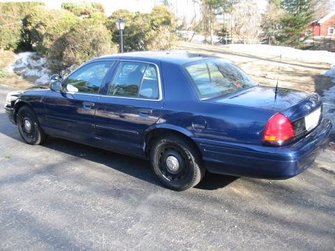 2003 Ford Crown Victoria - A dark blue 2003 Ford Crown Victoria sedan is parked on a paved surface with a wooded area in the background.
